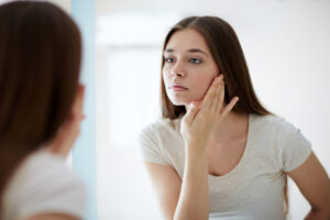 A woman examines her face in the mirror, considering whether she needs a facial in denver